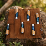 7-piece handmade kitchen knife set with patterned blades, wood and blue resin handles, and brown leather roll bag on natural log background
