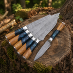 7-piece handmade kitchen knife set with patterned blades, wood and blue resin handles, and brown leather roll bag on natural log background