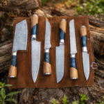 7-piece handmade kitchen knife set with patterned blades, wood and blue resin handles, and brown leather roll bag on natural log background